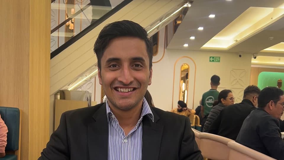 A headshot of Ayush Garg wearing a suit and shirt. He is smiling at the camera.